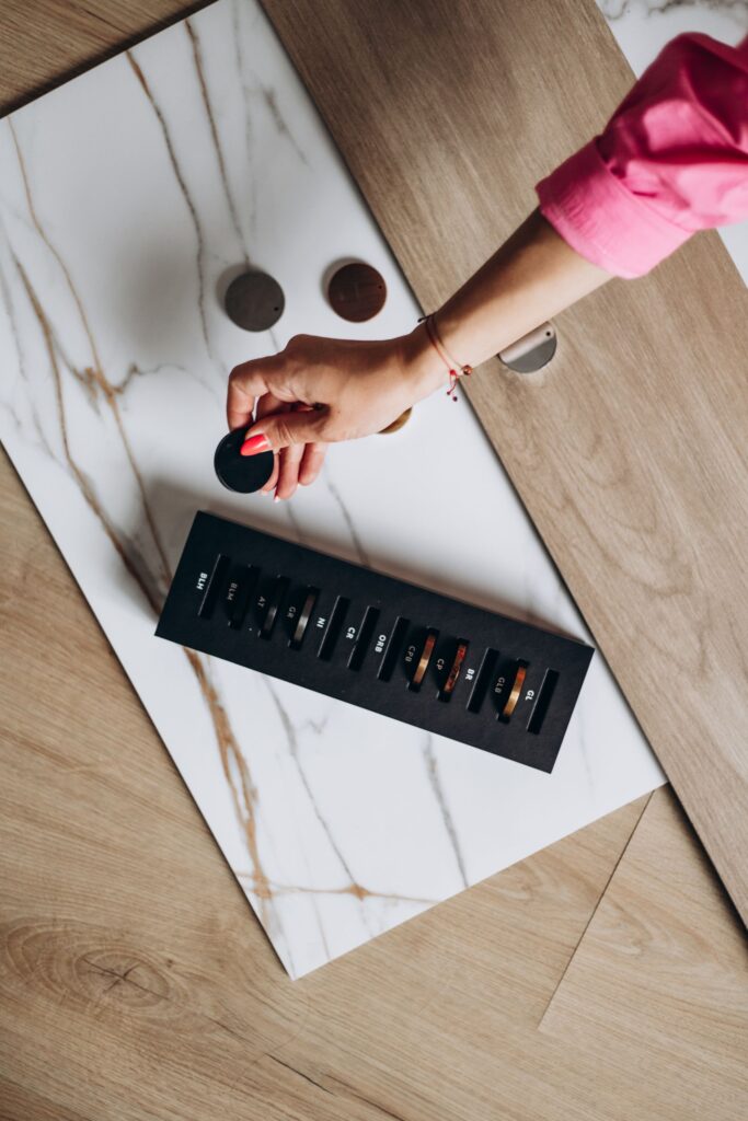 Ręka kobiety w różowej koszuli wybierająca okrągły próbnik koloru spośród próbek materiałów wykończeniowych; na stole leżą płyta imitująca marmur, panel drewniany oraz czarna paleta z próbkami kolorów i oznaczeniami.