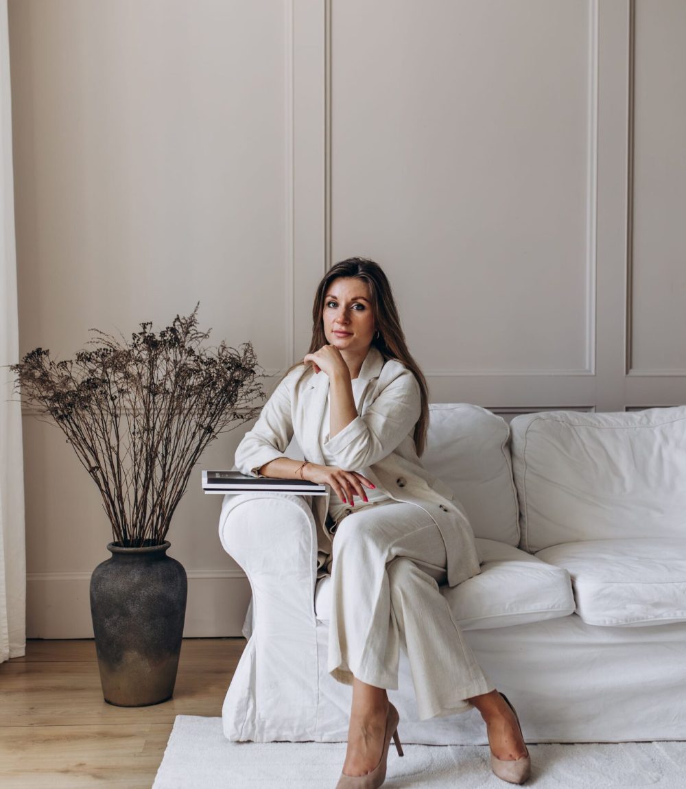 Na zdjęciu widoczna jest Ola Łabuda, architekt wnętrz, siedząca na białej sofie w elegancko urządzonym wnętrzu. Ubrana w jasny garnitur, trzyma w ręce książki o tematyce związanej z designem. W tle znajduje się duży wazon z suszonymi kwiatami oraz jasne ściany z delikatnymi panelami, które podkreślają minimalistyczny styl wnętrza.