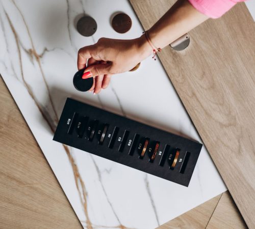 Ręka kobiety w różowej koszuli wybierająca okrągły próbnik koloru spośród próbek materiałów wykończeniowych; na stole leżą płyta imitująca marmur, panel drewniany oraz czarna paleta z próbkami kolorów i oznaczeniami.